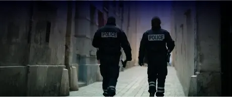 Two police officers in dark uniforms, with "POLICE" on their backs, walk down a narrow, dimly lit alleyway lined with tall buildings. One officer holds an object in their right hand. The setting suggests an urban environment, emphasizing themes of law enforcement and public safety.