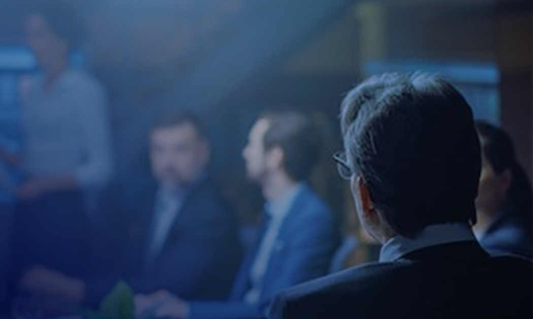 A meeting room with people in business attire, one person in focus from behind, others blurred in conversation.