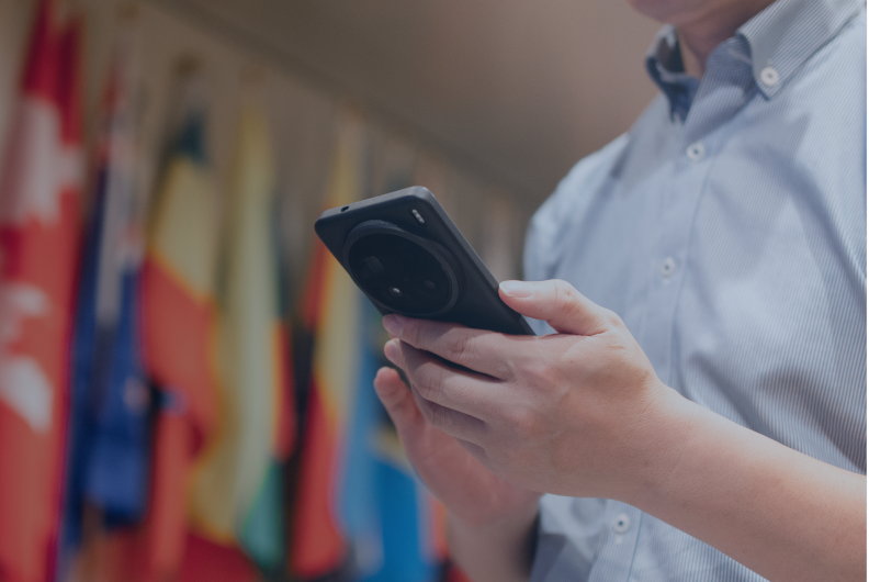 Person holding a mobile device using government-grade communications in front of blurred national flags; BlackBerry logo in corner.