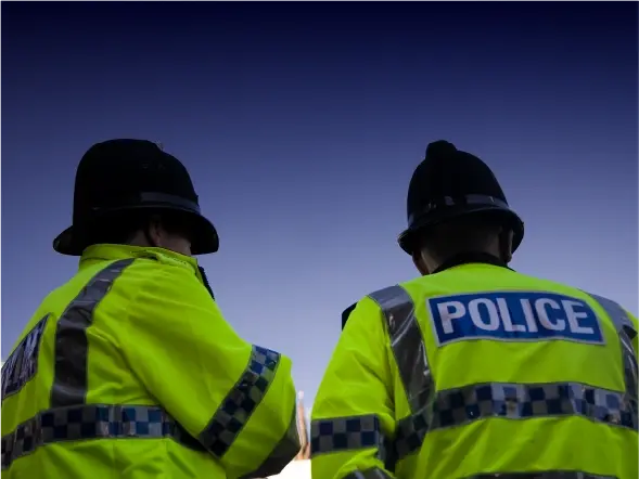 Two police officers in high-visibility yellow jackets with "POLICE" printed on the back, standing side by side. They are wearing dark helmets and facing away from the camera against a gradient sky background. The image emphasizes their roles in law enforcement during an outdoor event or patrol.