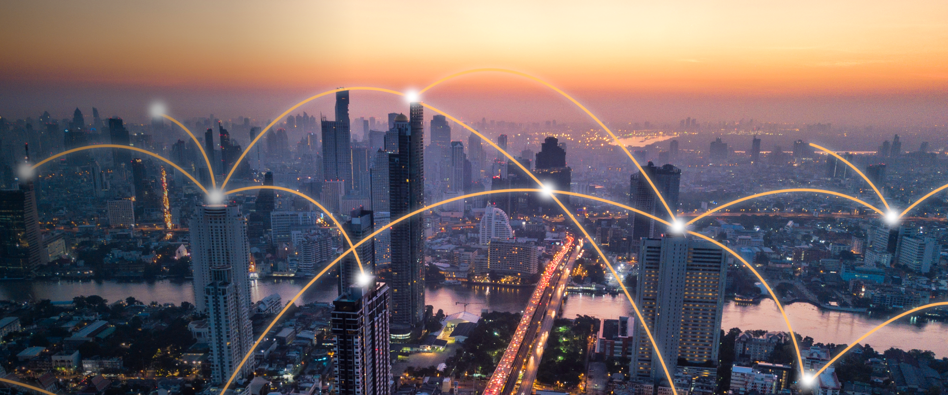 City skyline at twilight with skyscrapers connected by glowing arcs of light, symbolizing digital communication and connectivity. A river and highway run through the city, illuminated by warm hues of orange and pink in the sky.