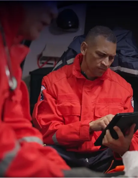 Two emergency personnel inside an ambulance. One is focused on a tablet, indicating engagement with digital information, while the other, slightly blurred, wears a stethoscope, suggesting a medical context. Their red uniforms with reflective stripes emphasize safety and visibility.
